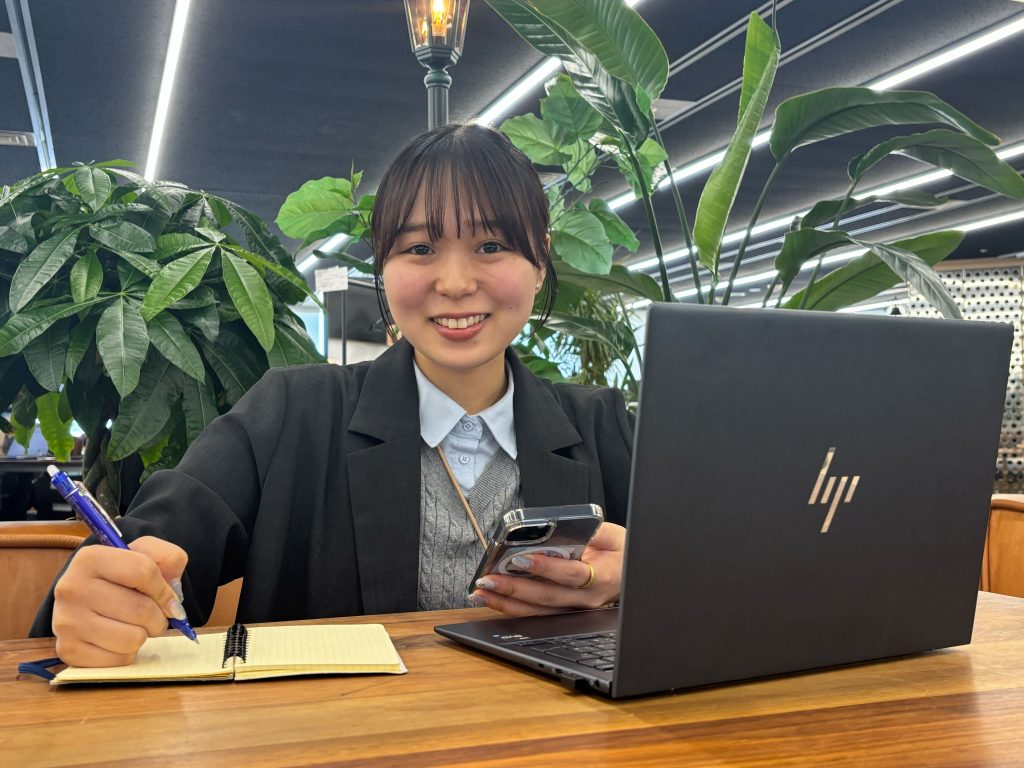 パソコンの前で、左手にスマホを持ち、右手でペンを持ち手帳を開いている女性の写真