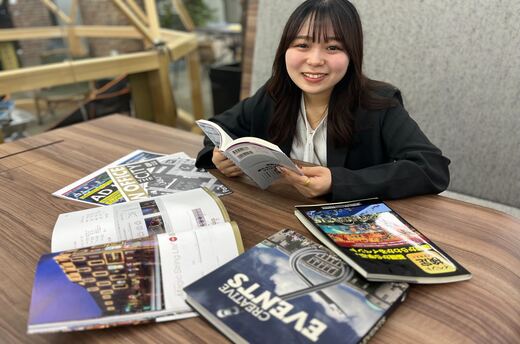 机の上に様々な本を並べ、本を手に持ちカメラ目線の女性
