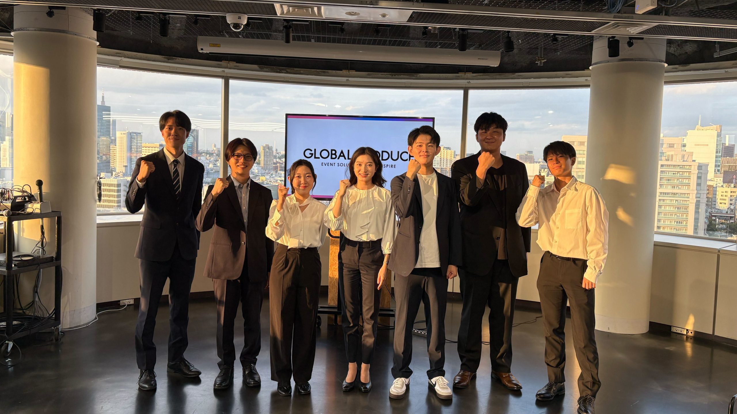 7人の内定者が楕円形の大会議室の中でガッツポーズをして、カメラ目線。