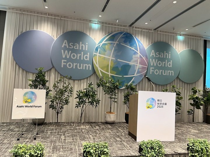 朝日地球会議(Asahi World Forum)のステージセット。白いカーテンを背景に、ブルー系の複数の円形パネルと、中央に大きな地球デザインのパネルが並んでいる。ステージ前にはロゴ入りの白い演台が置かれ、左右には観葉植物が多く配置されている。左側にはイベントロゴが印刷されたボードスタンドも見える。