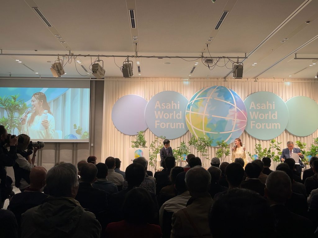 朝日地球会議の講演会場の様子。多数の聴衆がステージに向かって座っている。ステージ上には、中央に大きな地球儀のイラストと『Asahi World Forum』の文字がデザインされたパネルが設置されている。ステージ左側のスクリーンには、登壇者の一人であるゆうちゃみさんが映し出されており、彼女はマイボトルを持ってマイクに向かって話している。ステージには他に2名いる。