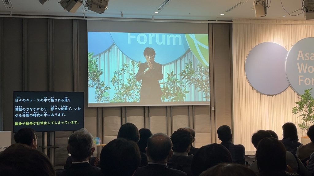 多数の聴衆が前方に注目している。前方の大型スクリーンには、手話通訳者が映し出されている。スクリーンの左下にあるモニターには演説内容のテキストが表示されている。