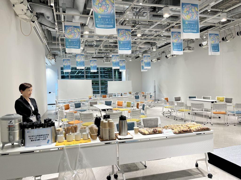 天井からイベントポスターが多数掲示された明るい会場内で、奥にはテーブルとカラフルな椅子が並べられている。手前にはコーヒーや軽食、ポットを並べたケータリングテーブルが設置されている。並んでいる紙コップは無漂泊の茶色のものである。ポッドの前には、「マイボトルをお持ちの方はウォータージャーから給水をお願いいたします」と書かれた札が置かれている。