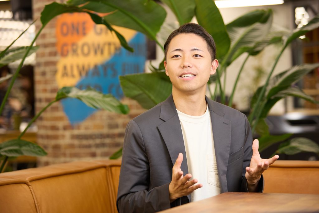 GLOBAL PRODUCEの社員がインタビューを受けています。カメラの奥のインタビュアーを見ながら両手を軽く開き、話しています。