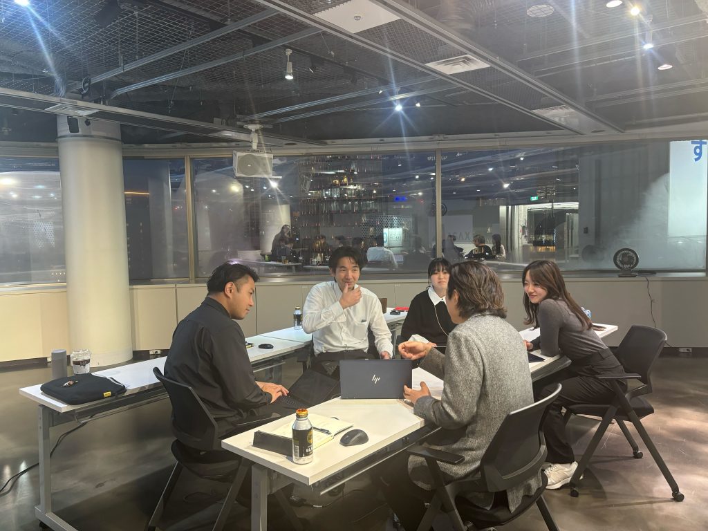 オフィス内の会議スペースで、複数名がテーブルを囲み、ノートパソコンを開きながら意見交換を行っている様子。参加者は向かい合って会話し、リラックスした雰囲気の中で活発なディスカッションが進められている。