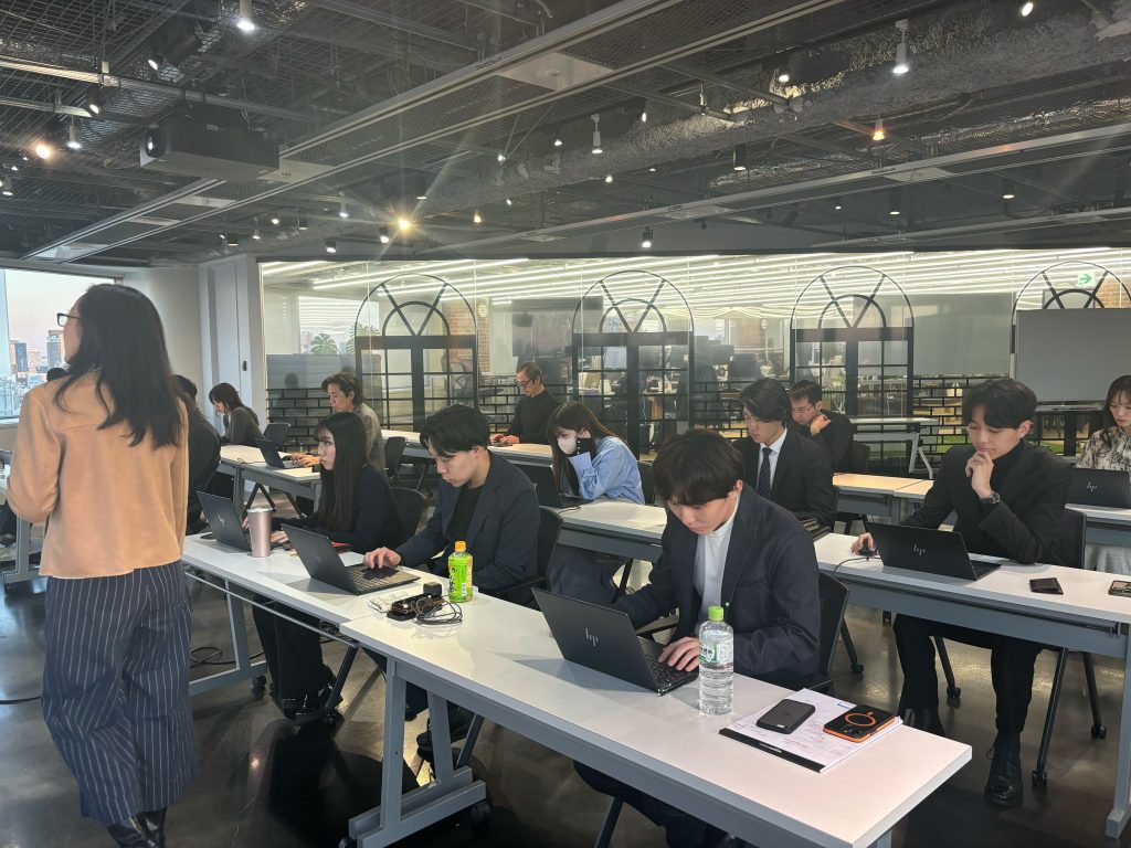 オフィス空間で、講師が前方に立ち、複数の参加者が長机に並んでノートパソコンを操作しながら研修を受けている様子。参加者はスーツ姿が多く、集中して作業に取り組んでいる。