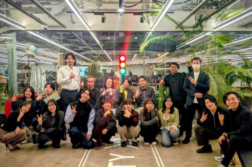 観葉植物が配置された開放的なオフィス空間で、複数名のメンバーが集まり、笑顔でポーズを取りながら撮影された集合写真。立っている人、しゃがんでいる人が混ざり、和やかで一体感のある雰囲気が伝わる。