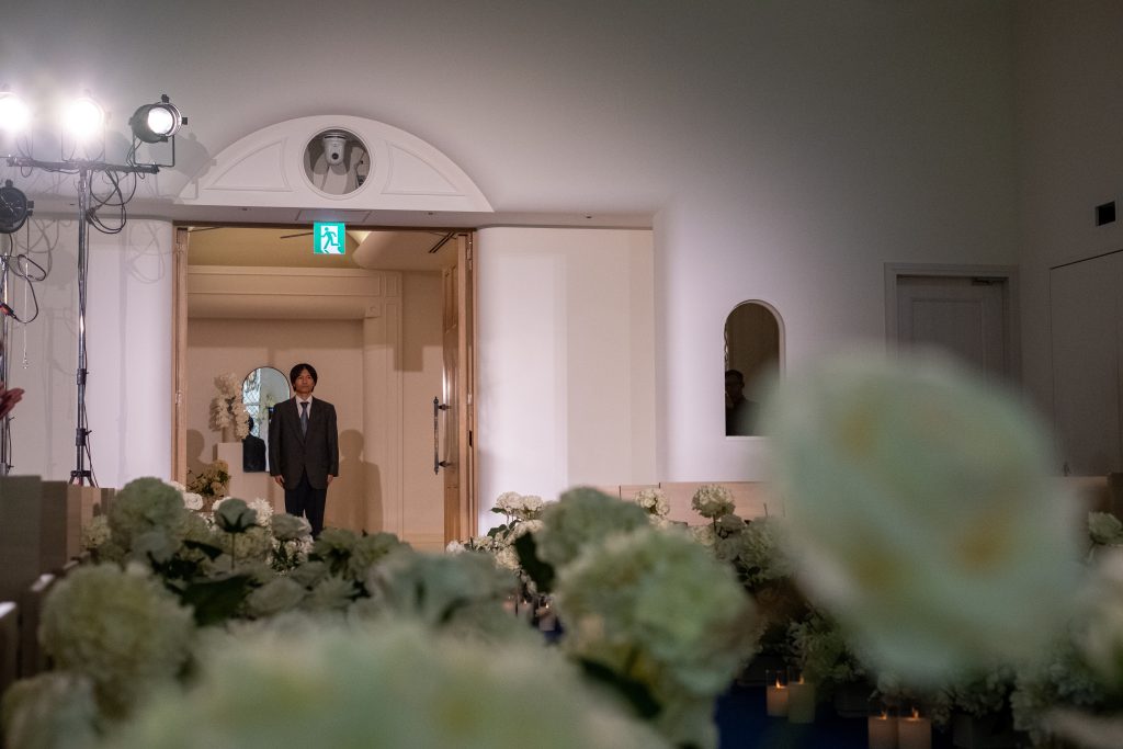 花で装飾された会場の扉から、スーツ姿の男性が入場してくる様子。
