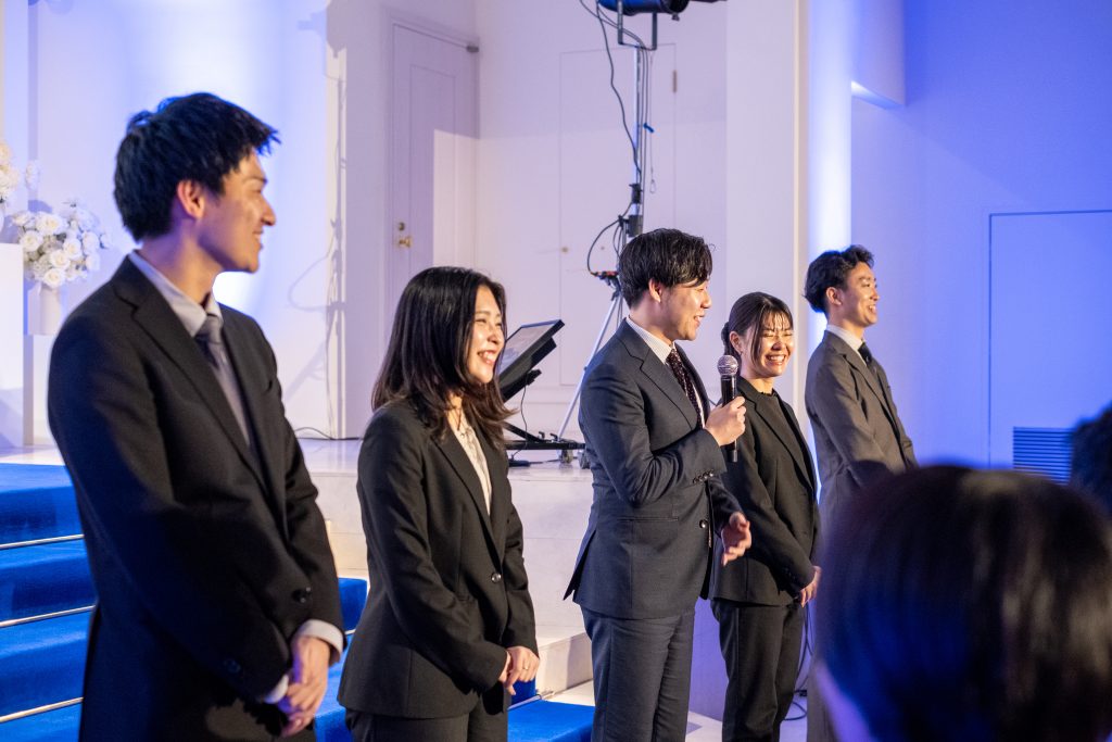 スーツ姿の男女がステージに並び、マイクを持った男性が話している様子。