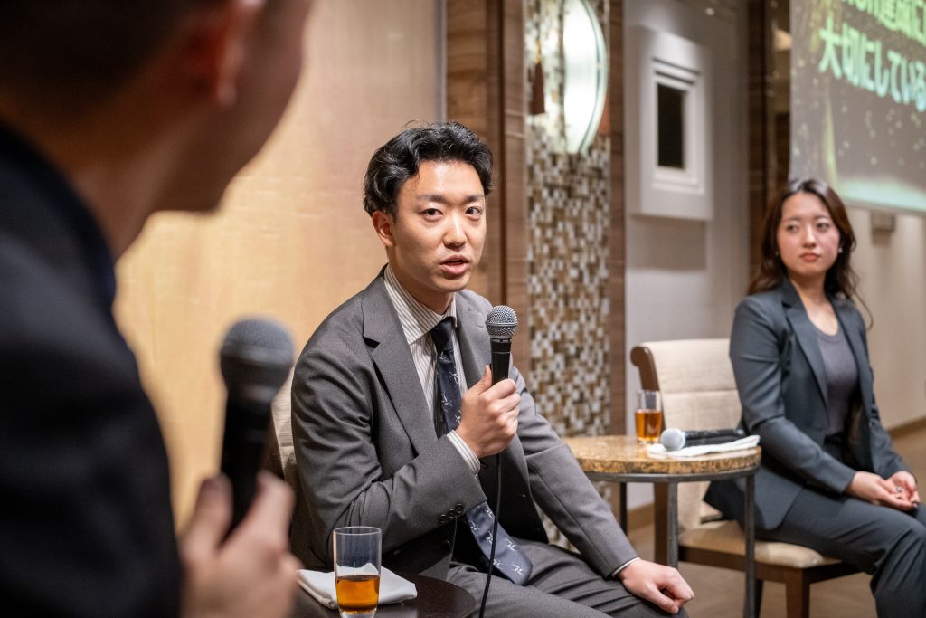 スーツ姿の男性がマイクを持って話し、隣で女性が話を聞いているパネルディスカッションの様子。