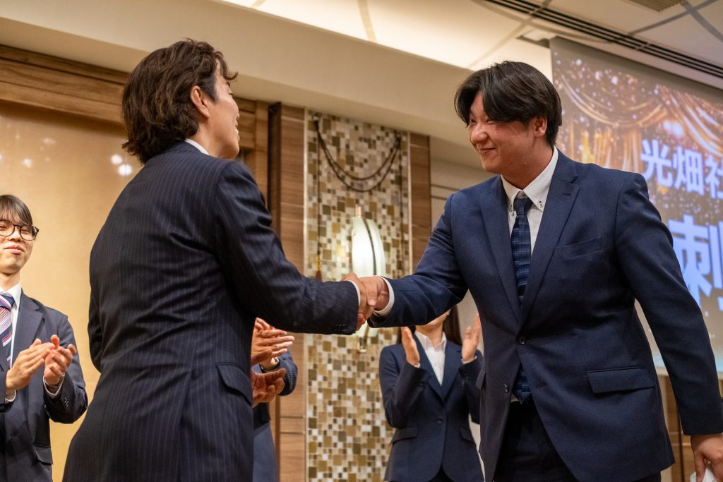 スーツ姿の男性2人がステージ上で握手を交わし、周囲の参加者が拍手している表彰シーン。