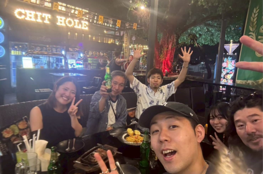 夜のオープンエアレストランで食事を囲み、乾杯やピースサインで盛り上がるグループのセルフィー。